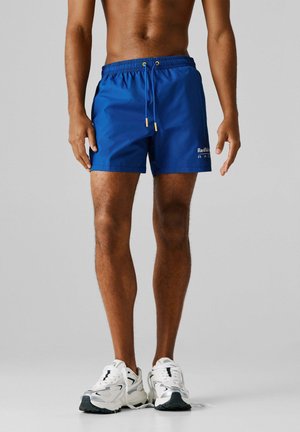 Torso e gambe maschili con pantaloncini blu Red Bull Racing e scarpe da ginnastica bianche, in piedi davanti a uno sfondo grigio uniforme.