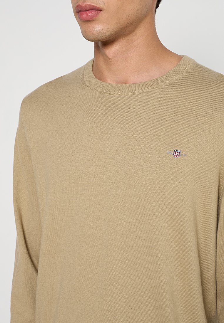 Beige Pullover mit langen Ärmeln und Rundhalsausschnitt, gerippten Bündchen und kleinem gesticktem Logo auf der Brust. Weiche Textur mit tailliertem Schnitt.