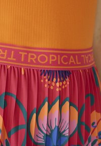 Top orange à côtelés avec une ceinture rose portant le texte "Tropical" ; en dessous, une jupe plissée aux motifs floraux vibrants avec des accents verts et bleus.