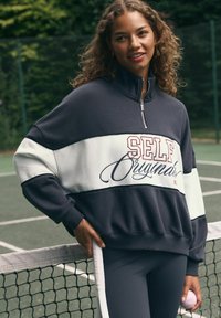 Tumehall ja valge lukuga dressipluus, millel on tekst "SELF Originals". Omab ribakaarte ja mugavat lõiget, kombineerituna hallide retuusidega tenniseväljakul.
