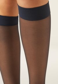 Průhledné černé podkolenky s tlustými neprůhlednými horními pásky, nošené na nohou na jednoduchém pozadí.