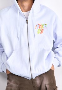 Lichtblauwe hoodie met ritssluiting gemaakt van zachte stof, voorzien van een voorvak en kleurrijke geborduurde tekst op de linkerborst.