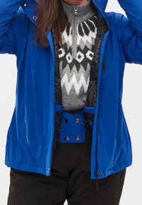 Chaqueta exterior azul con un alto cuello, cierre de cremallera y forro negro. Debajo, un suéter gris presenta un audaz patrón geométrico. Pantalones negros visibles.