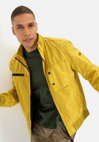 Gelbe, leichte Jacke mit einem Reißverschluss vorne, seitlichen Taschen und einer Brusttasche. Sie hat einen gerippten Kragen und ein kontrastierendes grünes Hemd darunter.