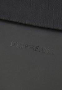 Fekete szövet sima tapintással; a felületen a "JOOP BEANS" dombornyomott logó található, letisztult, modern betűtípussal. Minimalista dizájn, túlnyomórészt sötét színárnyalat.