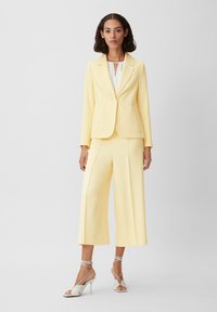 Yellow blazer with two pockets and a button closure, paired with wide-leg yellow pants. Model wears white strappy heels. Minimalistic design.