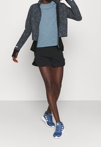 Černá a modrá vzorovaná zipová bunda, světle modré tričko, černé šortky a modré sportovní boty s zelenými akcenty.