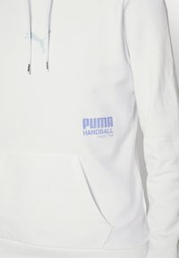 Sudadera blanca hecha de una tela suave, con un bolsillo tipo canguro. Logotipo de Puma en azul claro en el pecho y el texto PUMA HANDBALL en púrpura.