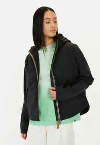 Zwarte, lichtgewicht jas met capuchon, voorzien van een gouden rits, twee voorzakken en elastische manchetten. Draag over een mintgroene sweatshirt.