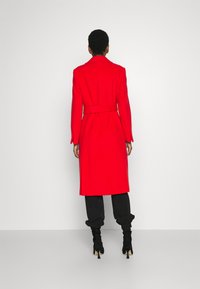 Roter, langärmeliger Trenchcoat mit einem Gürtel in der Taille, aus einem glatten Material gefertigt. Verfügt über einen Kragen und einen knielangen Schnitt. Über schwarzen Hosen getragen.