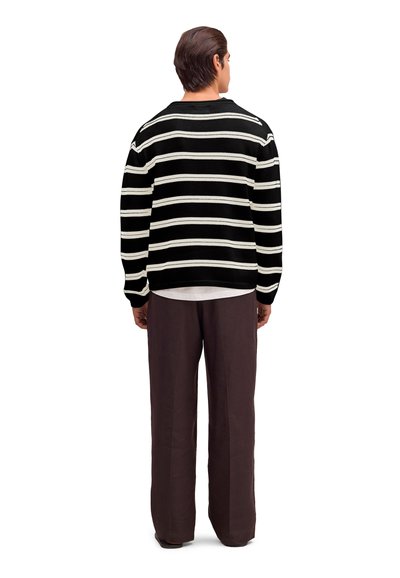 Mężczyzna stojący odwrócony tyłem, ubrany w czarno-biały sweter w paski, wystającą białą koszulę oraz luźne brązowe spodnie na białym tle.