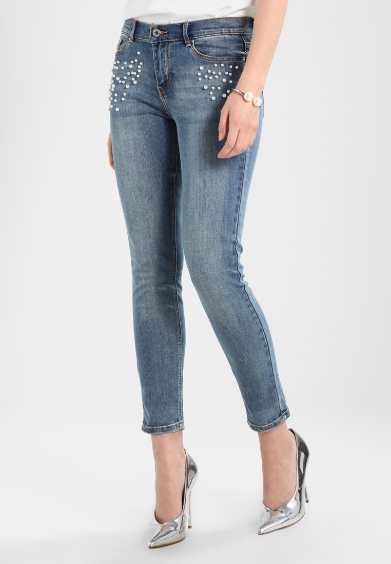Jean slim en denim bleu moyen, avec des ornements argentés sur les hanches, cinq poches et une taille mi-haute. Texture lisse.