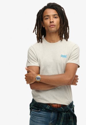 Light grey t-shirt with a round neckline and blue logo, paired with dark blue jeans. A gold watch and a plaid shirt tied at the waist.