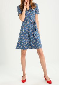 Blå blommig klänning med korta ärmar, omslutande midjeband och utsvängd nederdel. Tillsammans med röda högklackade skor.