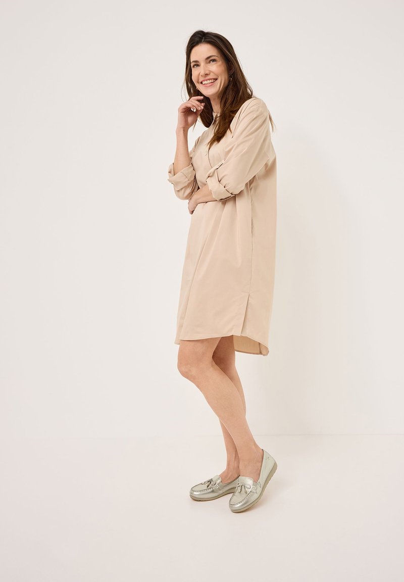Knælang beige skjortekjole med lange ærmer, knaplukning ved håndleddene, sidesplitter; båret med sølvfarvede loafers med sløjfe detalje.