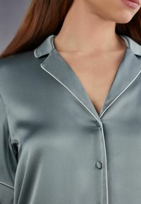 Silkig, dämpad teal pyjamaströja med klassisk krage, vit kantning och knappstängning vid bröstet, som visar upp en jämn textur.