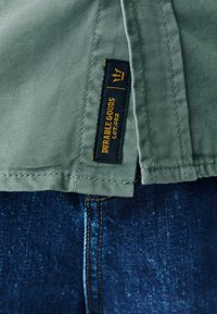 Grünes Stoffetikett mit "DURABLE GOODS" und einem Kronenlogo, auf ein strukturiertes Kleidungsstück genäht; darunter ist blauer Denim sichtbar.