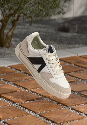 Hvid og beige sneaker med sorte logoaccents og snørebånd, hængende over trækvadrattæpper og små grå sten udendørs.