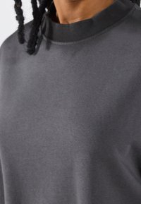 Primo piano di una persona che indossa una felpa grigia scuro con colletto a giro e cuciture visibili, tessuto a trama, capelli in dreadlocks neri.