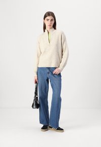 Beige ribbestrikket genser med glidelås til kvart, brukt over en grønn topp, kombinert med blå vide jeans og svarte sko, holder en svart håndveske.