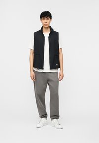 Νέος άνδρας που φοράει μαύρο γιλέκο με φερμουάρ, λευκή μπλούζα, γκρι φόρμα και λευκά αθλητικά παπούτσια, στέκεται μπροστά σε απλό λευκό φόντο.