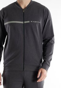 Graue Zip-Jacke mit strukturiertem Finish. Mit einem weißen gestreiften Design und Markenlogo auf der Vorderseite. Rippbündchen und Taillenbund.