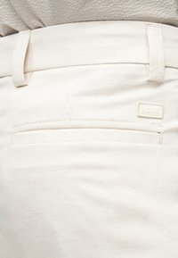 Κοντινή λήψη της μέσης μπεζ παντελονιού με θηλιές για ζώνη, οριζόντια πίσω τσέπη και μικρή ορθογώνια ετικέτα υφασμάτινου λογότυπου.