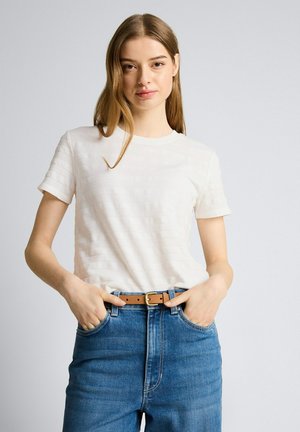 Młoda kobieta z długimi brązowymi włosami, ubrana w białą teksturowaną koszulę z krótkim rękawem, niebieskie dżinsy z wysokim stanem i brązowy pasek, ręce w kieszeniach.