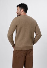 Beige herringbone-patterned sweater with ribbed cuffs and hem. Worn with brown pants, showing a relaxed fit and textured knit material.