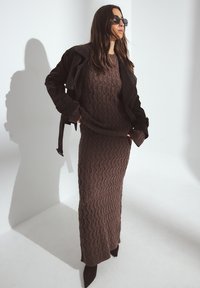 Brūns adīts kleita ar dimanta tekstūru, kārtota zem melna zamša žaketes. Melni norādīti augstpapēžu zābaki noslēdz izskatu. Ēna, kas krīt uz sienas.