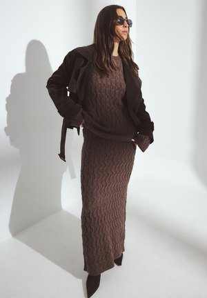 Vestido de punto marrón con textura de diamante, combinado con una chaqueta de ante negra. Tacones negros con punta completan el conjunto. Sombra proyectada en la pared.