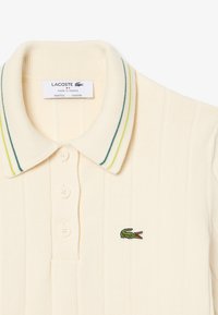 Polo a coste color crema con colletto, tre bottoni, dettagli a strisce verdi e gialle, e logo di coccodrillo verde sul petto.