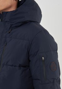 Giacca trapuntata blu navy con tasca sul petto con zip, cappuccio regolabile e patch del logo sulla manica sinistra. Presenta una texture liscia e trapuntata.