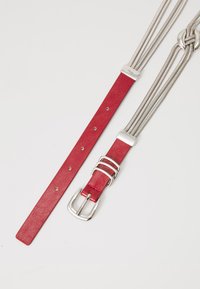 Rood leren riem met zilveren gesp en meerdere zilveren metalen kettingen bevestigd, gelegd op een witte achtergrond.