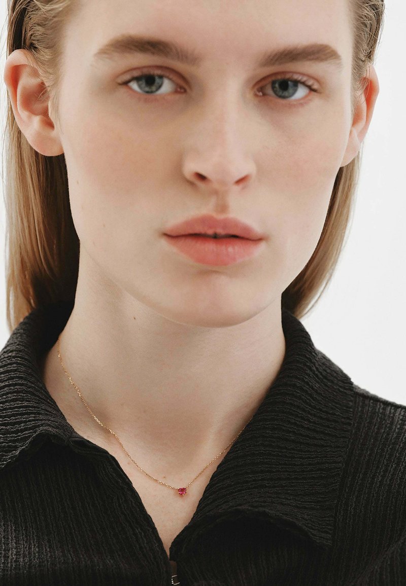 Collar de oro con una pequeña piedra preciosa roja, llevado sobre piel desnuda, contra un fondo de tela negra texturizada, resaltando los detalles de la joyería.