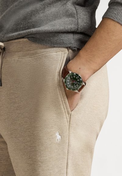 Grön urtavla med silverfärgad metall och brun läderrem. Beigea mjukisbyxor med en vit broderad logotyp. Grå tröja syns.