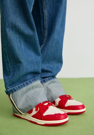 Jambes portant un jean bleu retroussé et des baskets rouge et blanc avec un design à lacets, debout sur un sol vert.