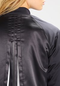 Svart bomberjacka med satinfinish, strukturerade paneler, ribbad krage och plisserad bakdetalj. Slät textur genom hela plagget.