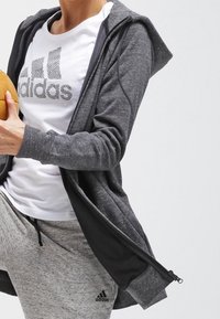 Persona vistiendo una camiseta blanca de Adidas, una sudadera con cremallera gris oscuro y pantalones de chándal grises claros de Adidas, sosteniendo un balón de baloncesto.