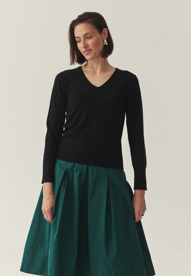 Pull noir à manches longues avec col en V, associé à une jupe plissée verte. La jupe présente une texture lisse et une silhouette ample.