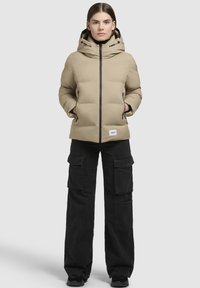 Beige Daunenjacke mit Kapuze, schwarzem Reißverschluss und seitlichen Taschen, kombiniert mit schwarzen Cargo-Hosen für einen kontrastierenden Look.