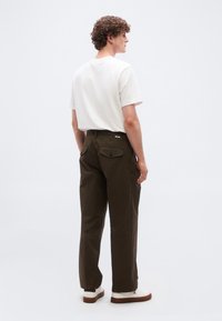 Νέος άνδρας με σγουρά μαλλιά που φοράει λευκό μπλουζάκι, σκουρόλαδι παντελόνι με πίσω τσέπες και λευκά αθλητικά παπούτσια σε απλό φόντο.