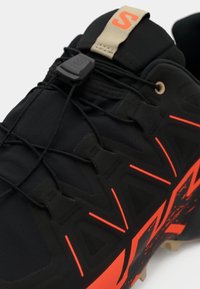 Scarpa sportiva nera con tomaia testurizzata, lacci elastici, dettagli arancioni vivaci e suola resistente con tacchetti multidirezionali.