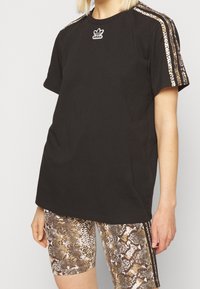 Musta puuvillainen t-paita, jossa on pyöreä pääntie ja lyhyet hihat. Olkapäillä on käärmekuvioiset yksityiskohdat, ja kauluksessa on Adidas-logo.