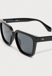 Μαύρα γυαλιά ηλίου με τετράγωνη κορνίζα και σκούρους φακούς, με διακριτό χρυσό λογότυπο στο μπράτσο, απεικονιζόμενα σε ανοιχτό φόντο.