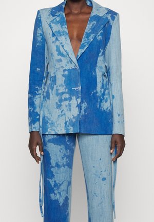 Personne à la peau foncée vêtue d'un costume délavé bicolore bleu avec un blazer à col en V profond et un pantalon assorti, debout devant un fond uni.