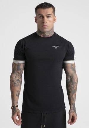 Mann med tatoveringer iført tettsittende svart SikSilk T-skjorte med logo på brystet og ermebånd, stående mot enkel lys bakgrunn.
