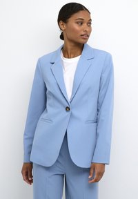 Světlomodrý blazer s klopami, zapínání na jeden knoflík, dvě přední kapsy a hladká tkanina, kombinovaný s bílým tričkem pod ním.
