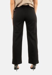 Pantalons larges noirs avec une taille élastique, texture lisse et coupe droite tombant jusqu'aux chevilles. Portés avec des chaussures à talons hauts noires.
