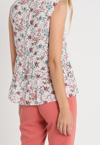 Květinová blůza v bílé barvě s růžovými a modrými vzory; bezrukávový design s nařaseným pasem; nošena s růžovými kalhotami, zobrazeno zezadu.